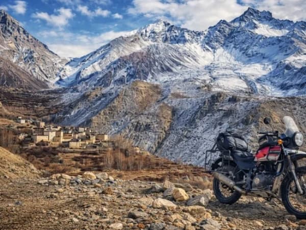 Adventure motorcycle parked on a rugged Himalayan mountain road with snow-capped peaks and a remote village in Nepal