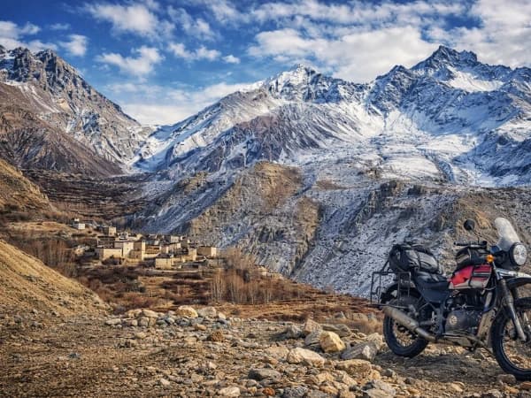 Adventure motorcycle parked on a rugged Himalayan mountain road with snow-capped peaks and a remote village in Nepal